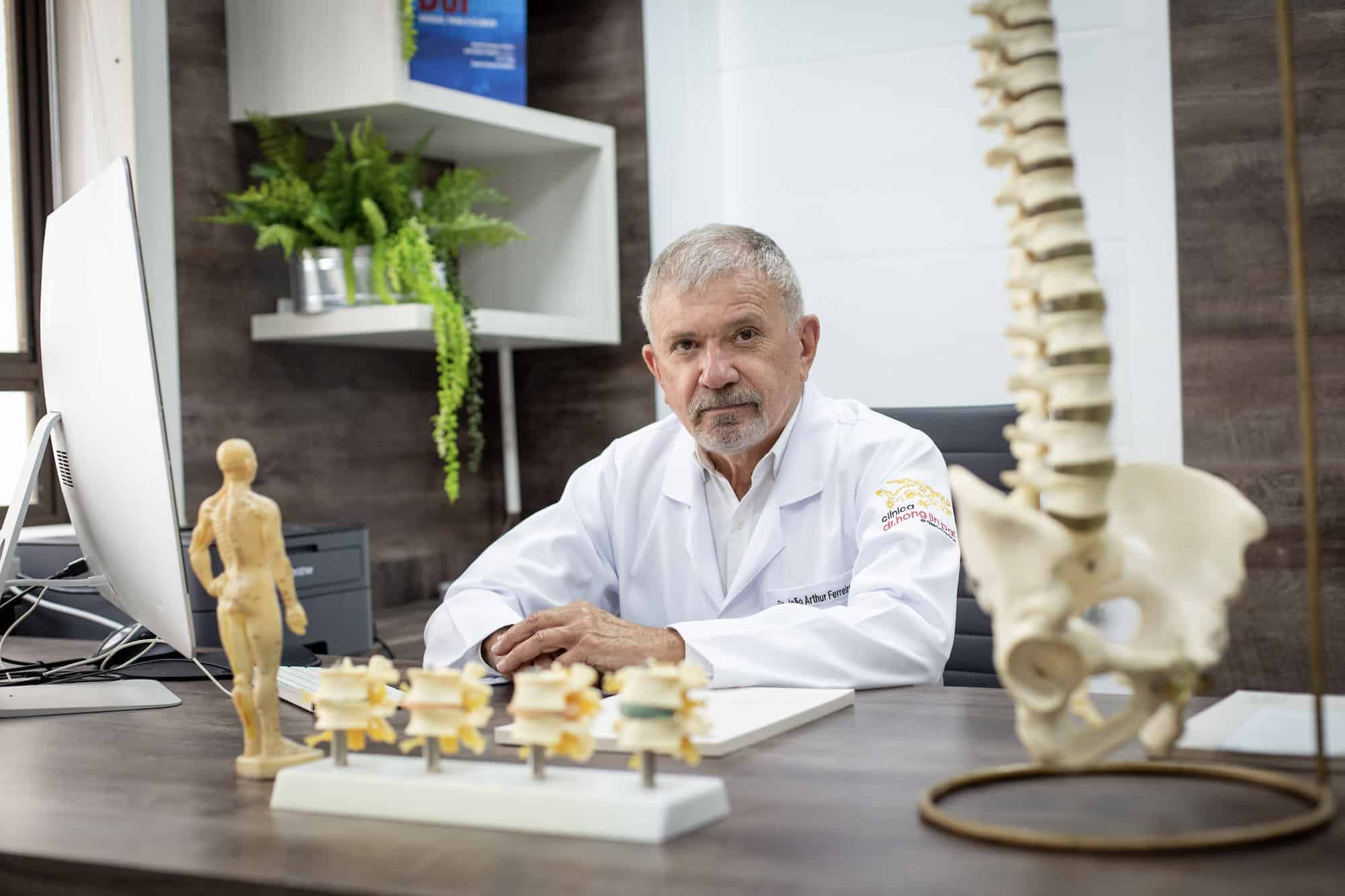 Dr. João Arthur Ferreira à mesa do consultório, com modelo de coluna.