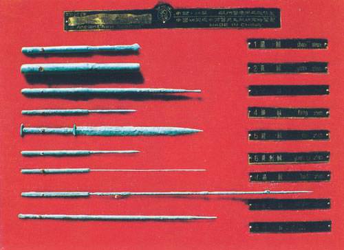 Conjunto antigo de agulhas de acupuntura de metal de diferentes espessuras e comprimentos fixado sobre uma placa vermelha com inscrições em chinês.