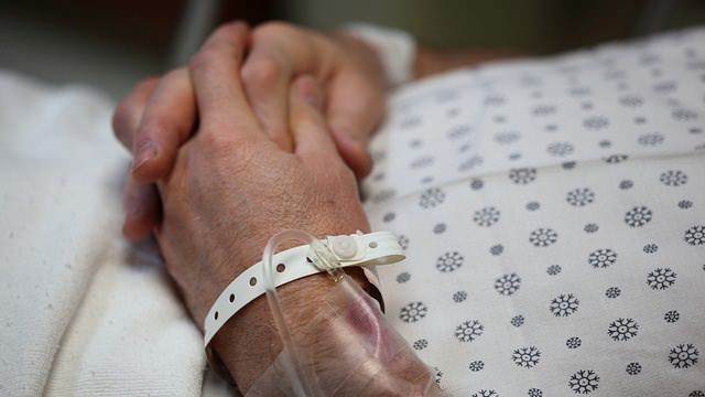 Mao de paciente idoso com pulseira de identificação hospitalar repousando sobre o braço, deitado em leito com avental hospitalar