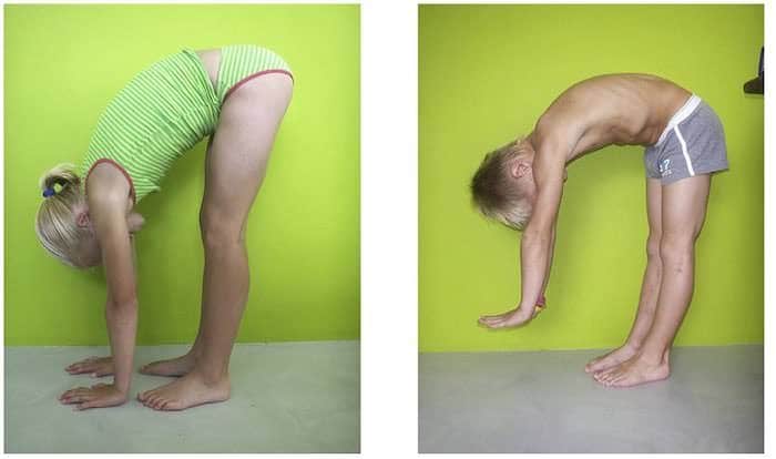 Duas pessoas em flexão do tronco com os joelhos esticados apoiando as palmas das mãos totalmente no chão, vistas de perfil contra parede verde.