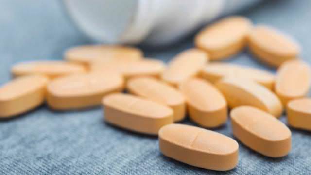 Comprimidos ovais alaranjados espalhados sobre tecido cinza ao lado de um frasco branco aberto.