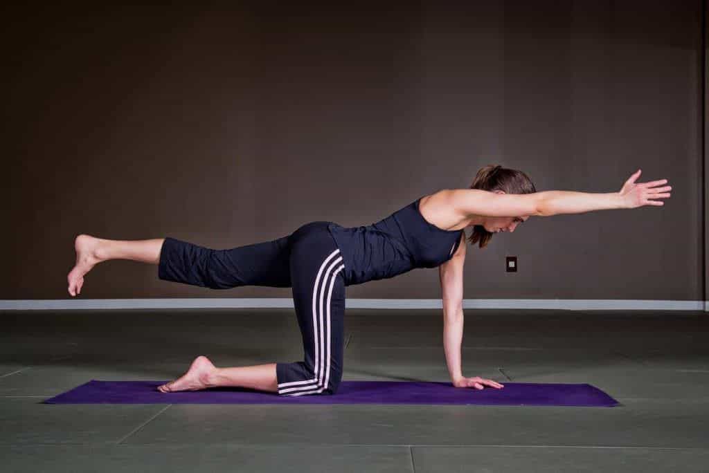 Mulher em roupa de ginástica executa o exercício de quatro apoios estendendo braço e perna opostos sobre um tapete de yoga.