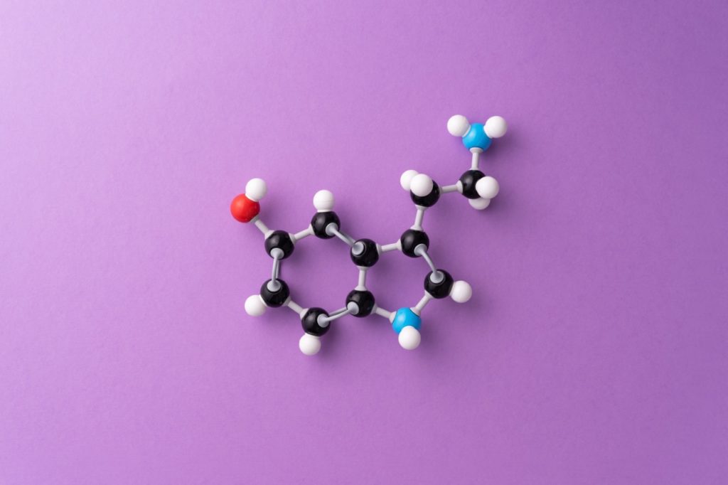 Modelo molecular tridimensional da serotonina em esferas pretas, brancas, vermelha e azuis sobre fundo lilás