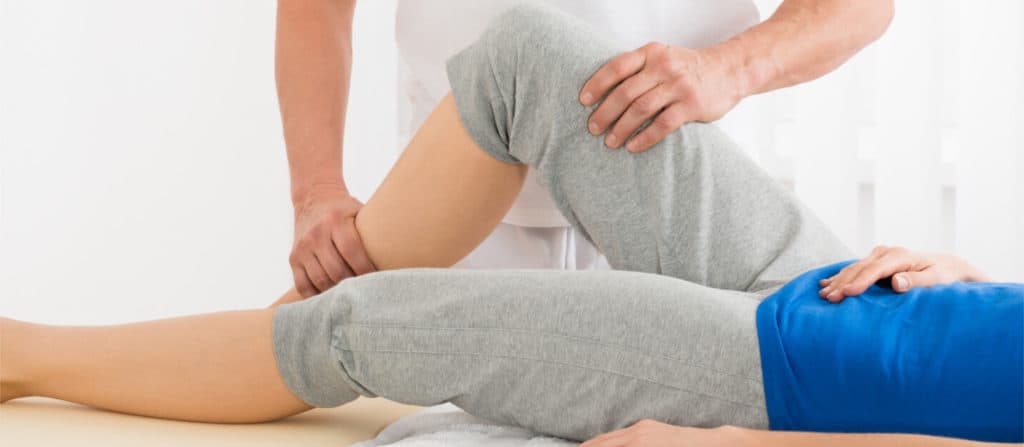 Profissional mobilizando o joelho flexionado de uma pessoa deitada na maca durante sessão de fisioterapia.