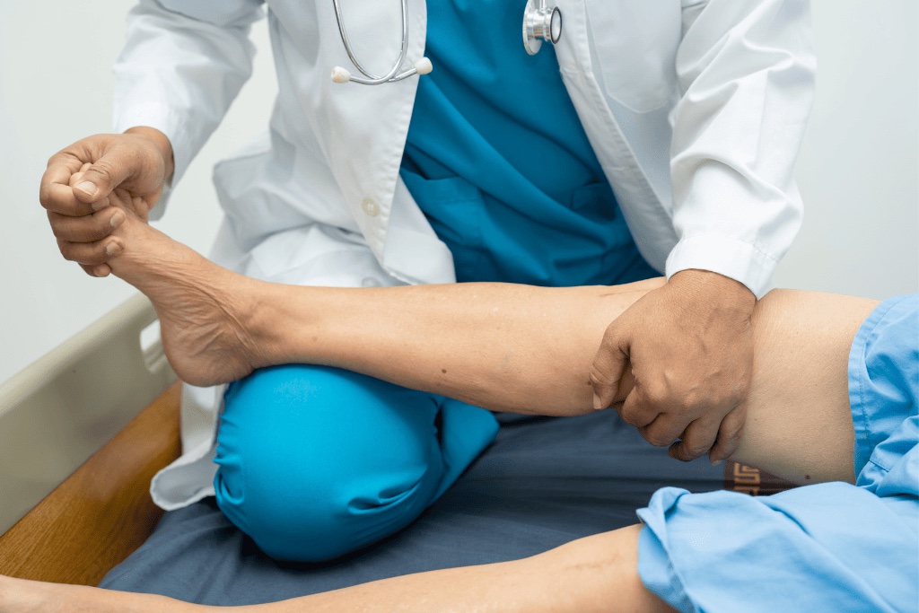 Médico de jaleco branco com estetoscópio examinando a perna de um paciente deitado em maca de consultório.