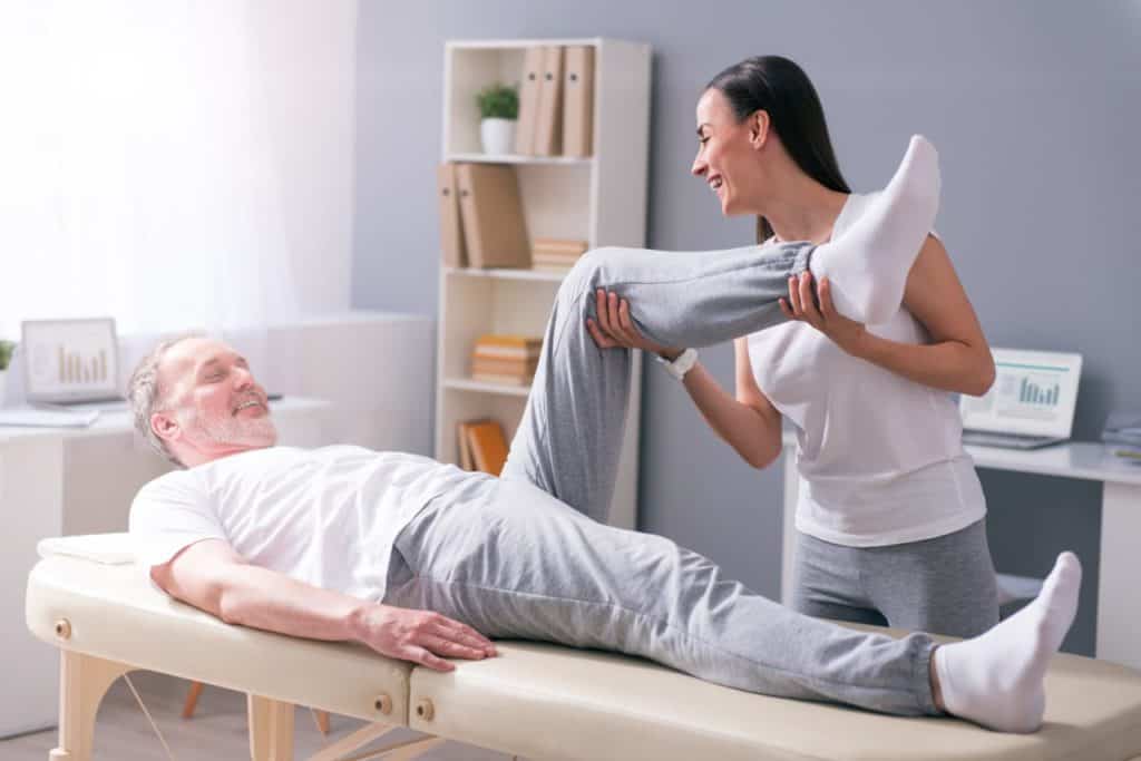 Fisioterapeuta alongando a perna flexionada de um paciente idoso deitado em uma maca durante a sessão.