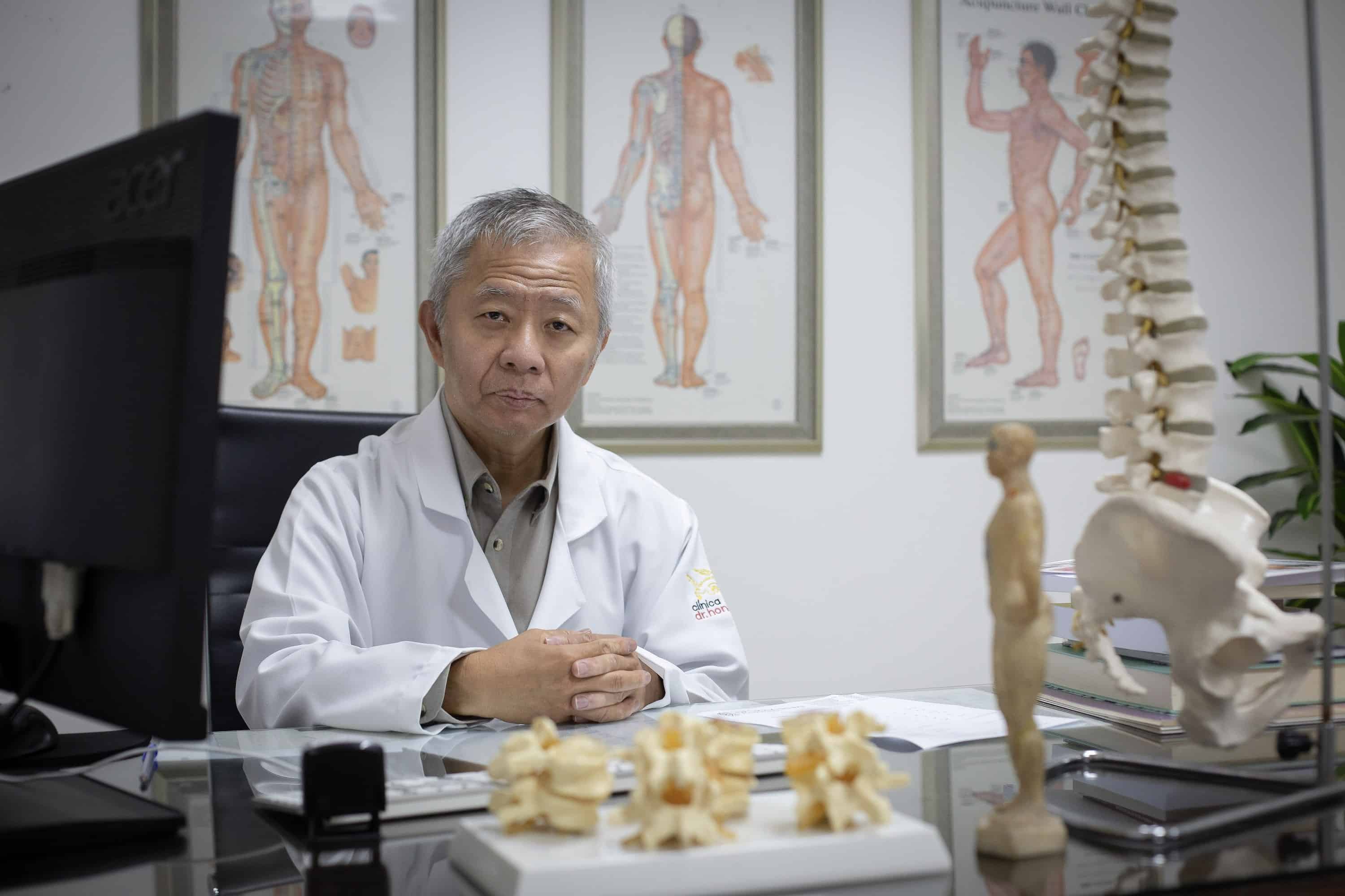 Prof. Dr. Hong Jin Pai na mesa de atendimento, com mapas de acupuntura ao fundo