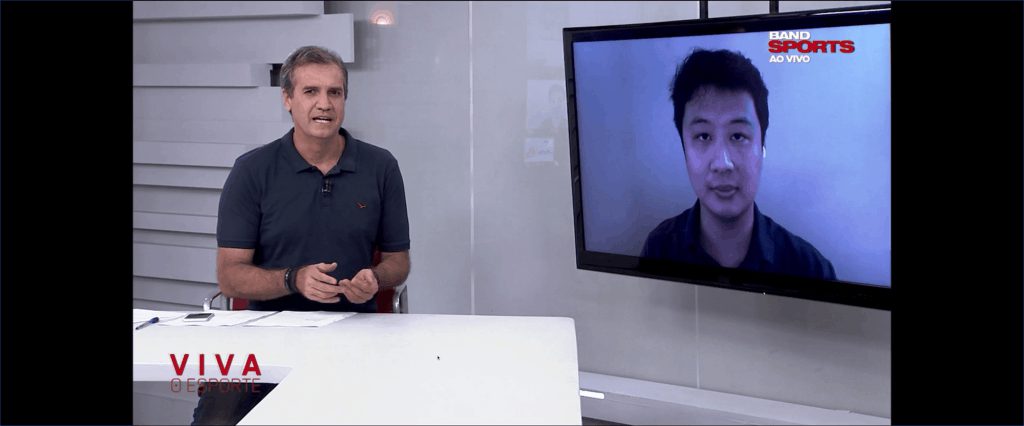 Apresentador em bancada de estúdio; telão exibe médico por videochamada com marcas BandSports ao vivo e Viva o Esporte