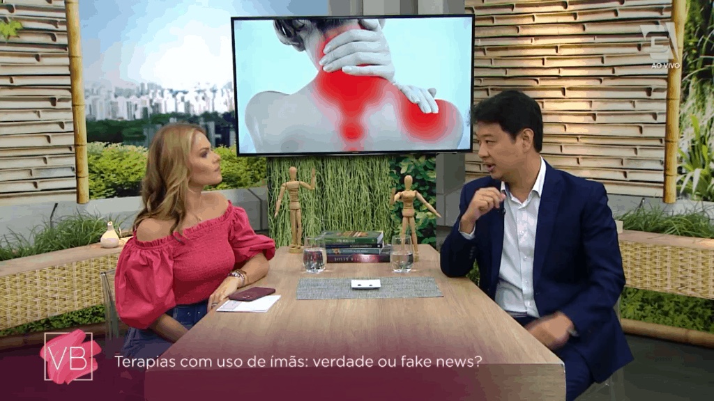 Apresentadora e médico à mesa; telão mostra pescoço e ombro com áreas vermelhas e letreiro sobre terapias com ímãs
