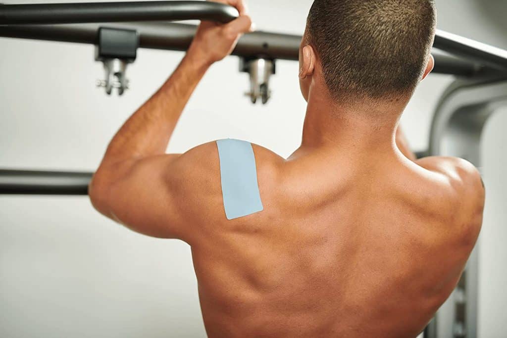 Homem sem camisa, de costas, com um adesivo retangular azul colado sobre o músculo do ombro/trapézio em uma academia.