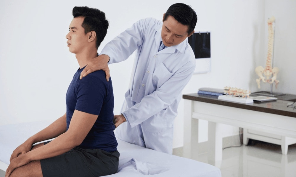 Médico de jaleco examina as costas de um paciente sentado em uma maca, com modelo de coluna ao fundo no consultório.