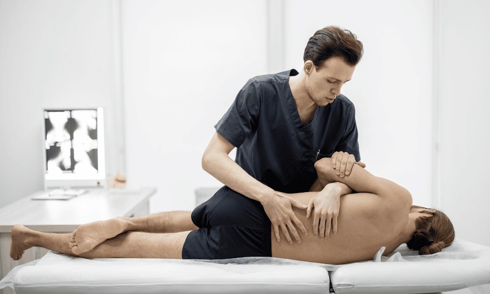 Profissional realiza técnica de terapia manual na coluna de paciente deitado de lado na maca, com negatoscopio exibindo radiografia ao fundo.