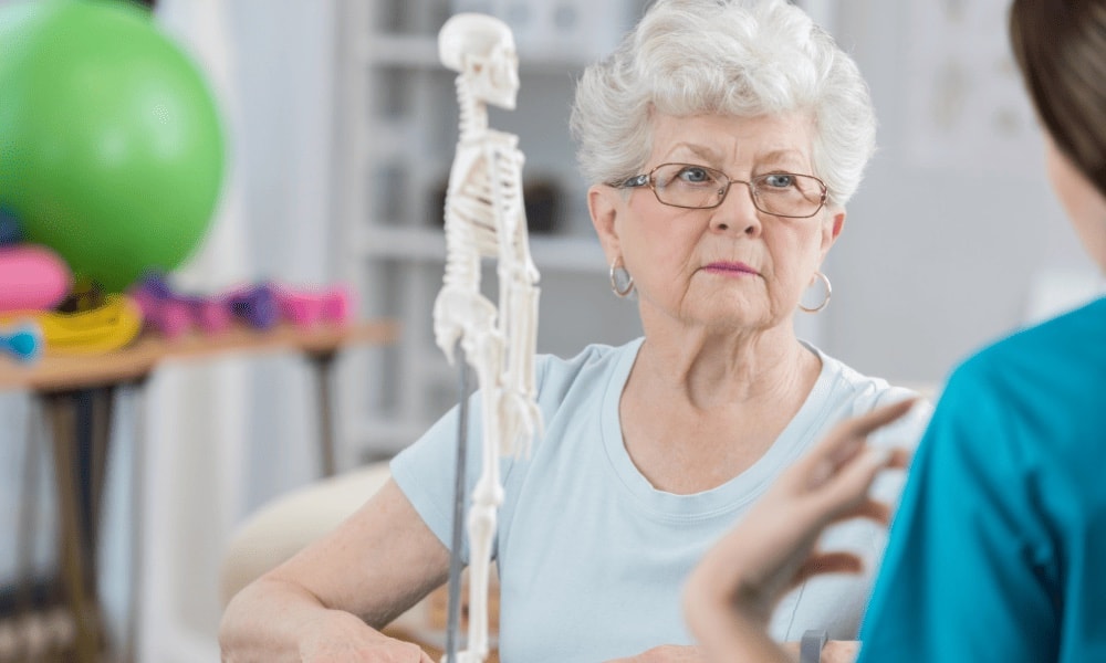 Mulher idosa sentada em sala de reabilitação, com modelo de esqueleto, bola de exercício, faixas elásticas e halteres ao fundo, conversando com uma profissional de saúde.