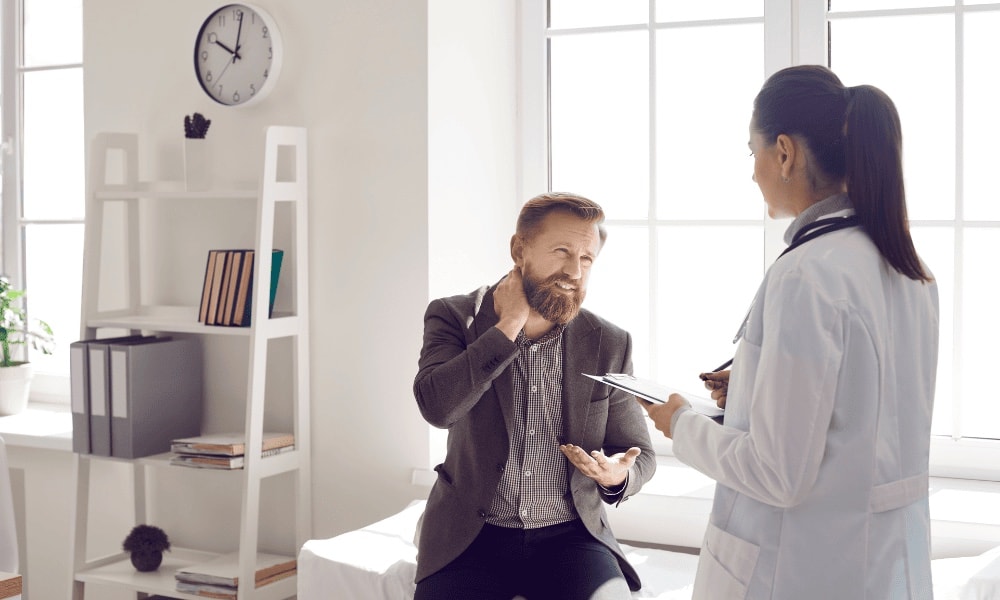 Medica de jaleco branco com prancheta conversando com um paciente sentado que toca o próprio pescoço, em um consultório claro.