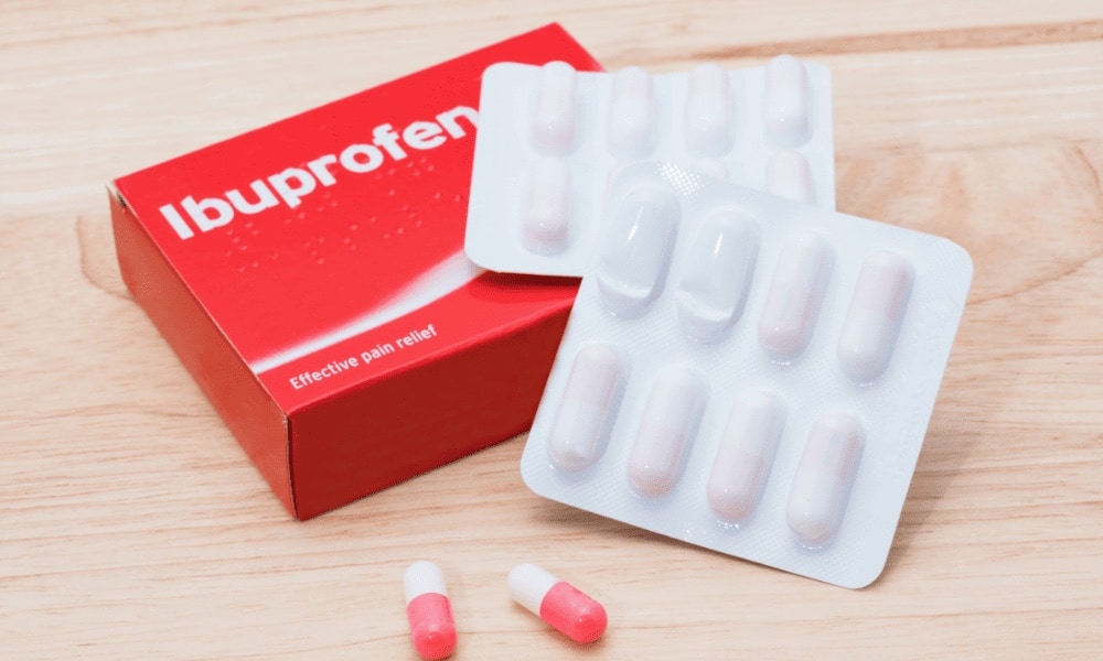 Caixa vermelha de ibuprofeno ao lado de uma cartela de comprimidos e duas cápsulas rosa e branca sobre superfície de madeira