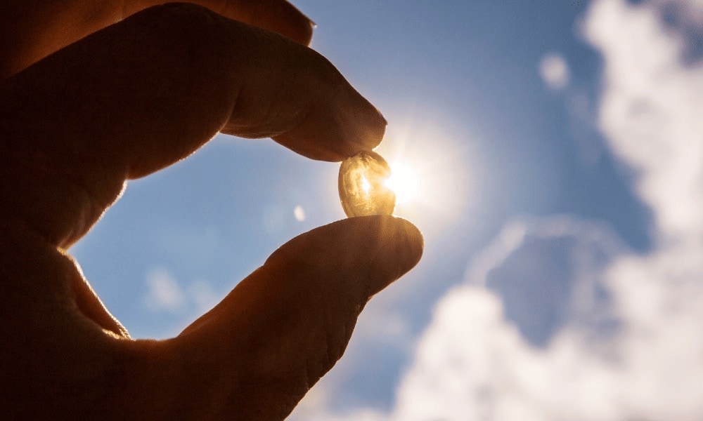 Mao segurando uma cápsula de vitamina D contra o ceu azul, com o sol brilhando atrás da cápsula