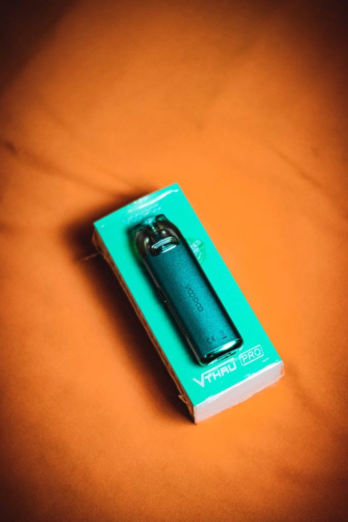 Dispositivo de vape (cigarro eletrônico) dentro da embalagem verde apoiado sobre tecido alaranjado.
