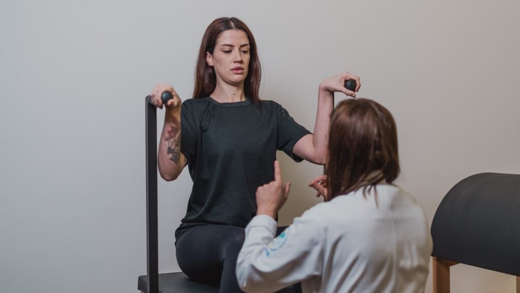 Paciente sentada segura pequenos pesos com os braços enquanto a fisioterapeuta, de costas, conduz o movimento de frente.