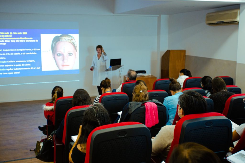 Medica ministrando aula de acupuntura com slide de pontos para plateia em auditorio