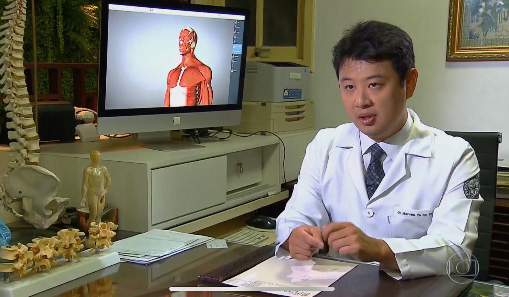 Dr. Marcus Yu Bin Pai de jaleco em entrevista de TV na mesa com modelo de anatomia muscular ao fundo