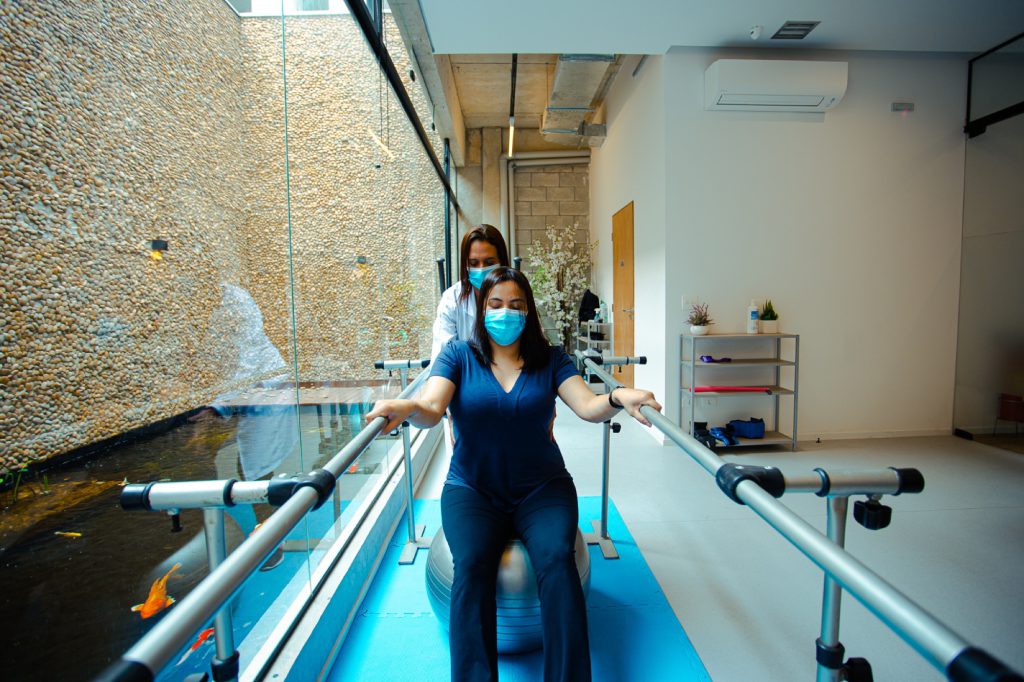 Fisioterapeuta supervisionando paciente sentada em bola de exercício entre barras paralelas