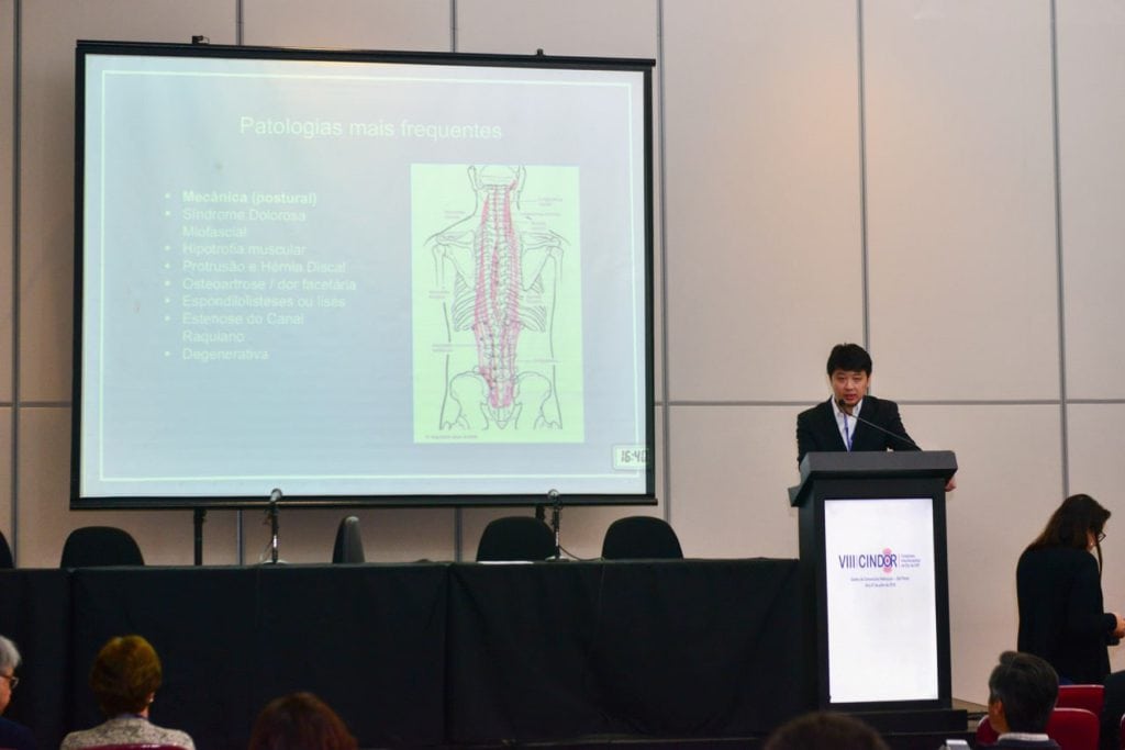 Dr. Marcus Yu Bin Pai ministrando palestra no congresso VIII CINDOR ao lado de slide sobre patologias da coluna