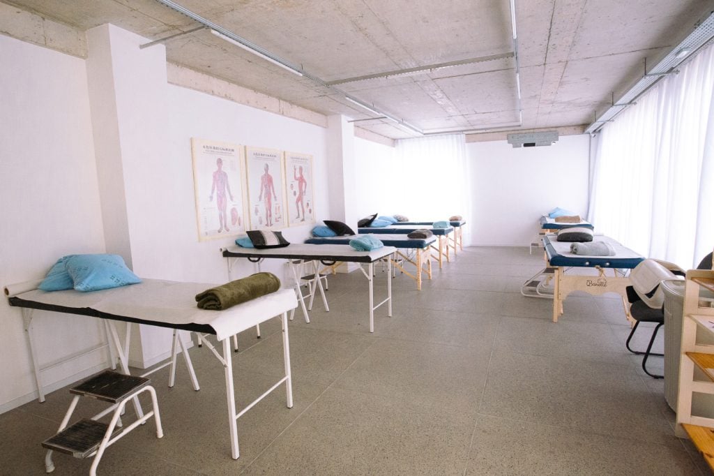 Sala coletiva de tratamento com várias macas de acupuntura e posters anatômicos na parede