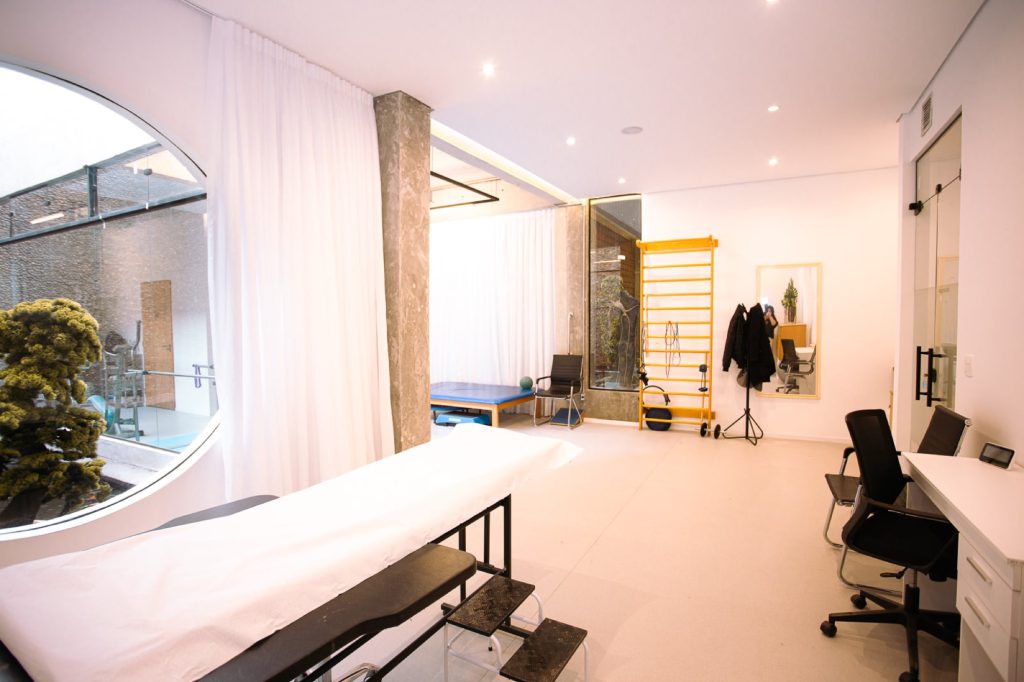 Dor no psoas e iliopsoas: lombar, virilha e quadril 2 Sala de fisioterapia iluminada com maca, espaldar amarelo e janela redonda para o jardim