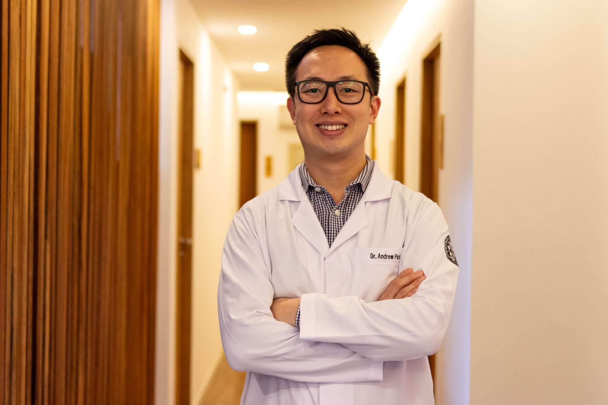 Dr. Andrew Park de jaleco branco, braços cruzados, no corredor das salas de tratamento.