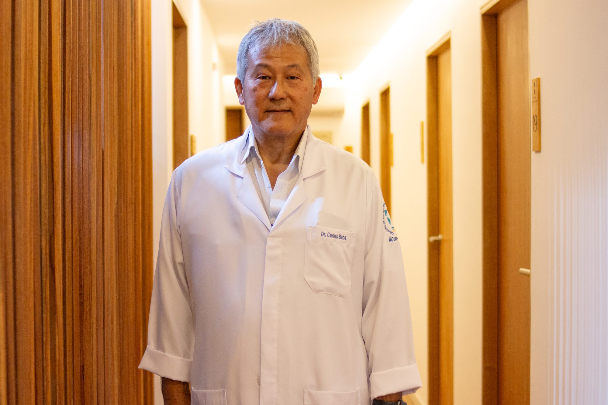 Dr. Carlos Roberto Babá de jaleco branco no corredor das salas de tratamento da clínica.